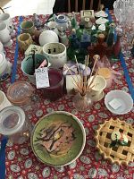 Overview of bottles, candles, green ceramic bowl, ceramic apple pie dish, and other kitchenware items arranged on a patterned tablecloth