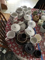 Overview of table with multiple ceramic items including white covered soup bowls, various decorated mugs and brown ware pottery on red floral tablecloth.