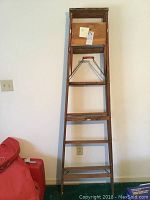Full front view of wooden step ladder standing against white wall with red bag nearby.