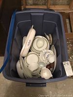 Blue plastic storage bin containing assorted Corelle dishware including plates, bowls, cups, gravy boat, butter dish, and shakers.