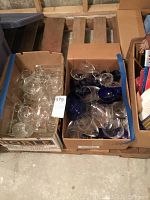 Two cardboard boxes filled with mixed clear and cobalt blue glassware including tumblers, brandy snifters, wine glasses, and small bowls.