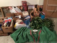 Two green artificial Christmas trees with tree skirts, boxes of ornaments, gift bags, wicker baskets, and greenery decoration in basement setting.