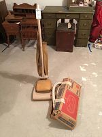 Full view of the vintage Hoover upright vacuum cleaner standing upright with flexible hose coiled beside it and an original cardboard box for attachments placed nearby.