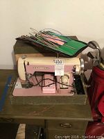 Full view of vintage pink Brother sewing machine with attached foot pedal and control knobs, sitting on top of green carrying case with visible wear.