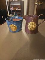 Purple watering can and blue bucket candle side by side showing sunflower appliques