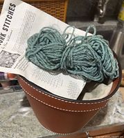 Needlepoint instructions sheet, green yarn skein, brown faux-leather basket