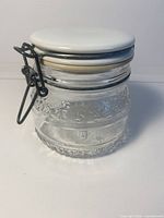 Side view of clear embossed glass canister with attached white ceramic lid and wire bale