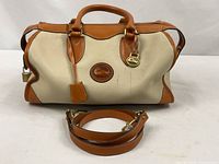 Front view of satchel showing cream pebbled leather, tan trim, rolled handles, brass duck fob, detachable strap placed in front