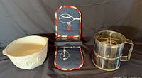 Group shot of cream ceramic mixing bowl, blue double oven glove folded, and metal flour sifter