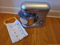 Frigidaire stand mixer with stainless steel bowl, splash guard, and white chef apron displayed on wooden floor