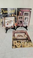 Front covers of three cross-stitch pattern books on stands