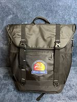 Front view showing top flap, buckle straps, front zip pocket, mesh sides, promotional sticker