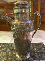 Front view of silver-plated cocktail shaker showing handle, spout and grapevine band