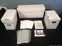 Three white comic storage boxes, stacks of bags and boards on black surface