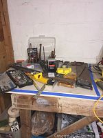Photo showing assorted hand tools on wooden workbench against a white block wall, includes saws, chisels, crowbars, measuring tape, hammer, and other hand tools.