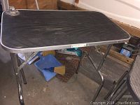 Photo of General Cage dog grooming table with black ribbed top and foldable metal legs on a concrete floor.
