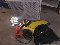 Photo shows two animal life jackets in yellow and orange on the floor, a folded white plastic security gate behind them, and other items.