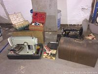 Two sewing machines side by side. Left is Kenmore model 1120 in light gray on a black base, right is black vintage Every Ready machine with wooden case beside it. Several woven baskets filled with sewing supplies behind the machines.