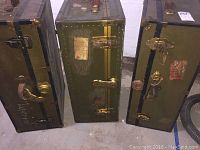 Three vertically standing olive-green travel trunks with brass hardware and black trim, showing visible wear and age marks on a concrete floor.
