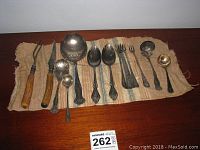 Full layout of all silver plated utensils on cloth runner, showing forks, spoons, ladle and serving pieces