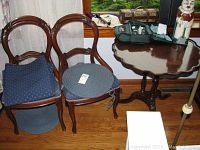 Side view of 2 wooden side chairs with blue upholstered cushions and the round wooden side table with pie crust edge in the background