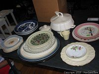 Photo showing a collection of assorted plates, soup tureen, and drinking glasses on a table.