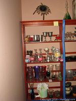 Full shelf view showing stainless steel cups, goblets, glass candle holders, vases, and assorted candles arranged on four wooden shelves. Bottom shelf includes flower pot with artificial flowers.