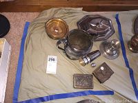 Overview of silver plated decorative items including water pitcher, cigarette case, trinket box, covered dish, bowl, candlestick holder and small cup.