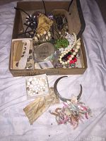 Box containing assorted costume jewelry including beaded necklaces, metal necklaces, earrings, and shell necklace along with a silver-tone engraved metal headband and rhinestone chain necklace laid out beside it.