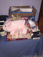 Overview photo of boxed items including board games Scrabble and Monopoly, several stuffed animals including a large pink pig and smaller rabbits, various card games and boxed DVDs spread on a table.