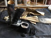 Photo showing English riding boots, helmet, riding crop, and black leather chaps arranged on black floor covering.