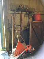 Wide view of barn wall with four yard rakes, two shovels, tamper, red bucket on shelf, and orange leaf blower on floor.