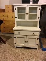 Front view of white painted child's wooden cabinet with glass doors and two lower doors, showing brass hardware and overall wear.
