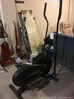 Full view of the black OrbiTrek elliptical machine with foot pedals visible, showing its size and surrounding basement environment with other items.