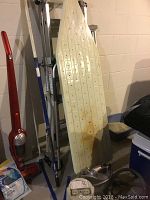 Photo showing red and white Conair clothing steamer leaning against the wall beside metal ironing board with rust stains.