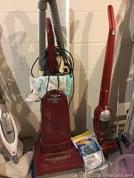 Full lot showing two red vacuum cleaners side by side; one upright and one stick style with accessories and vacuum bags.