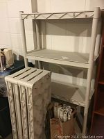 Three-quarter view of assembled plastic shelving unit with three shelves, beige color, next to unassembled shelves stacked vertically.