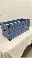 Angled view of blue wood crate showing slatted sides, black metal corner brackets, and side handle