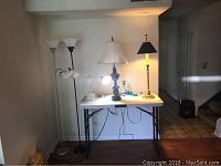 Six lamps: two tall white torchiere floor lamps, two table lamps (one with white shade and ornate base, one with black shade and striped base) on a white folding table
