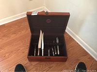 Open wooden box containing stainless steel serving utensils with ornate handles arranged inside.
