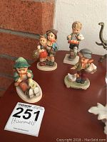 Four Hummel figurines on display with lot number visible, showing variety of poses and sizes.