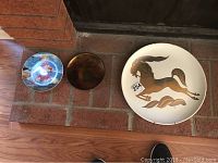 Three decorative plates arranged on brick surface. Left plate depicts child at beach, center smaller copper-toned decorative plate, right larger plate with stylized horse design.