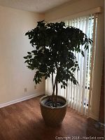 Full view of the faux Ficus tree standing in the ceramic pot inside a room with hardwood flooring and window blinds.