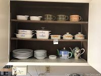 Full view of shelving with Corningware dishes, soup mugs, white plates, bowls, and two decorative teapots on the bottom right.