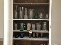 Full view of various glassware on three shelves inside a wooden cabinet including clear glass vases, green vases, and various glass drinking vessels.