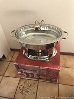 Oval stainless steel chafing dish with glass lid placed on its original box, showing its polished finish and side handles in a tiled corner.