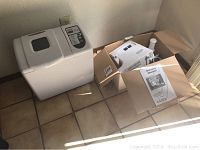 Overall view of white Chef's Mark bread maker and boxed meat grinder food processor with owner's manual on tiled floor