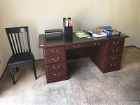 Wood laminate desk with multiple drawers and brass handles, dark finish, visible damage in lower corner, with items on top and a dark wood chair with black padded seat beside it.