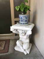 Full view of white concrete cherub statue holding a platform with a blue and white ceramic pot containing a green plant.