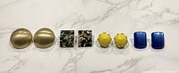 All four pairs of clip-on earrings laid out in order: brass round, black/gold rectangle, yellow dome, blue rectangle